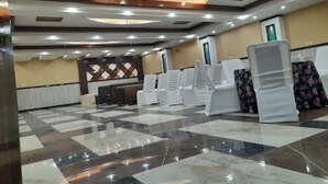 A spacious hall with a checkered floor, white chairs, and a reception area with a decorative wall. - Hotel Amba Ambience (Hathras)
