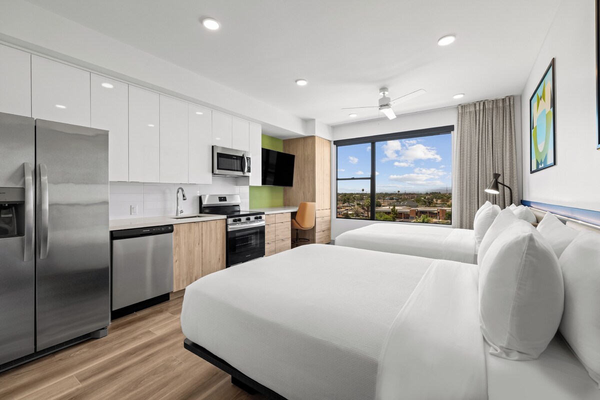 A modern hotel room with a kitchen, a large bed, a view of the city, and a ceiling fan.