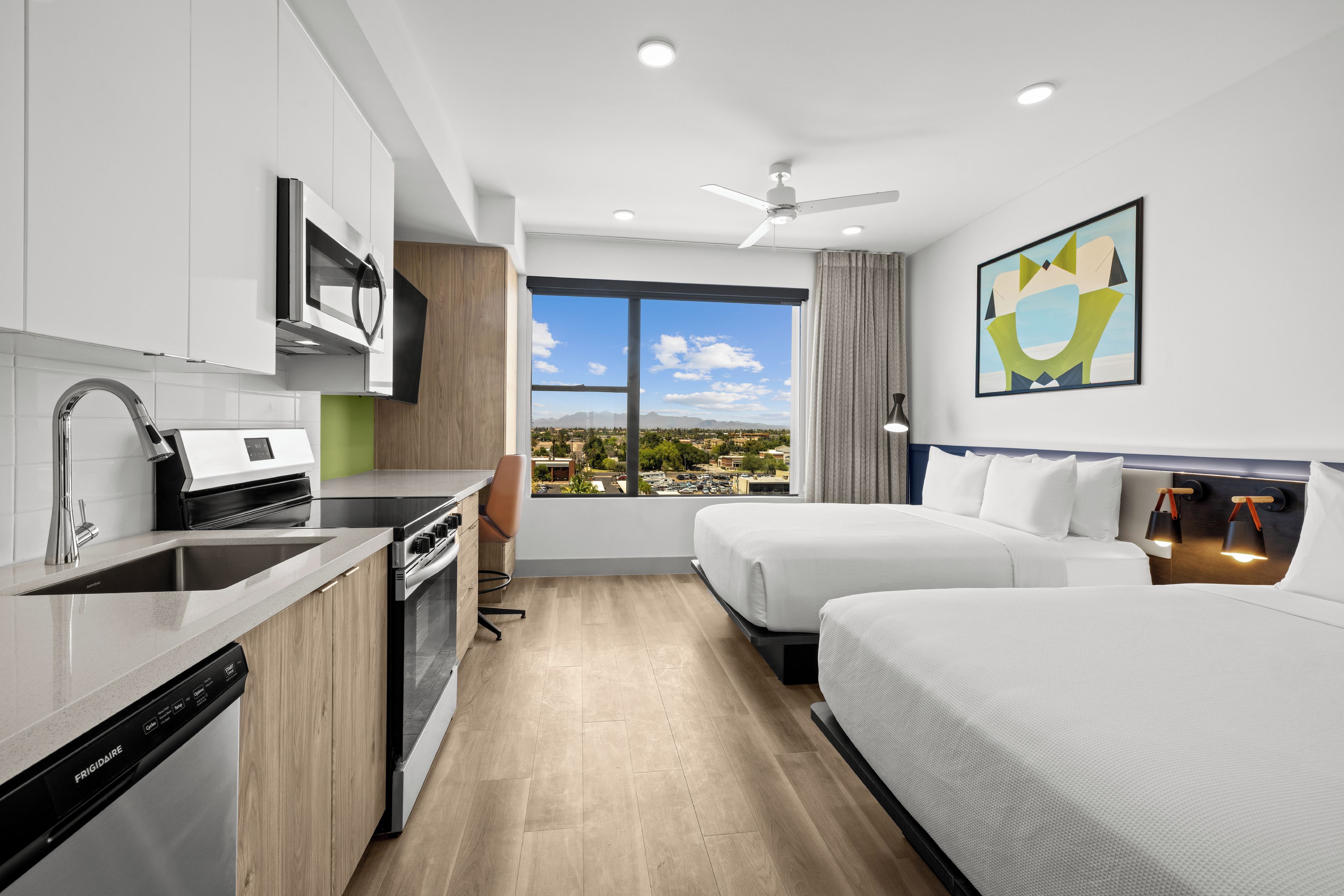 Una habitación de hotel moderna con kitchenette, un gran ventanal con vista a la ciudad, cama y ventilador de techo.
