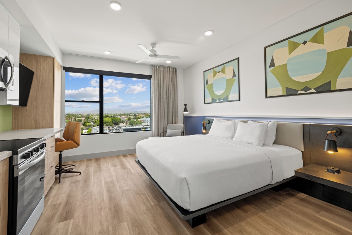 A modern hotel room with a large bed, a desk, a chair, a view of the city, and artwork on the walls.