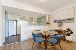 A modern kitchen with white cabinets, a central island, and blue chairs around a glass table. - The Beaufort Street Nook (Perth)