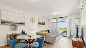 A modern living room with a round glass table, blue chairs, a beige sofa, and a large window offering a view of the outdoors. - The Beaufort Street Nook (Perth)