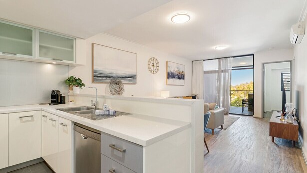 A modern kitchen with white cabinetry, a built-in dishwasher, and a large island with a sink. There is a dining area with a table and chairs, and a view of the outdoors through large windows. - The Beaufort Street Nook (Perth)