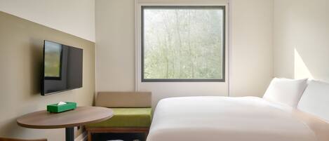 Une chambre d’hôtel avec un lit, une télévision, une petite table, un banc et une fenêtre donnant sur des arbres.