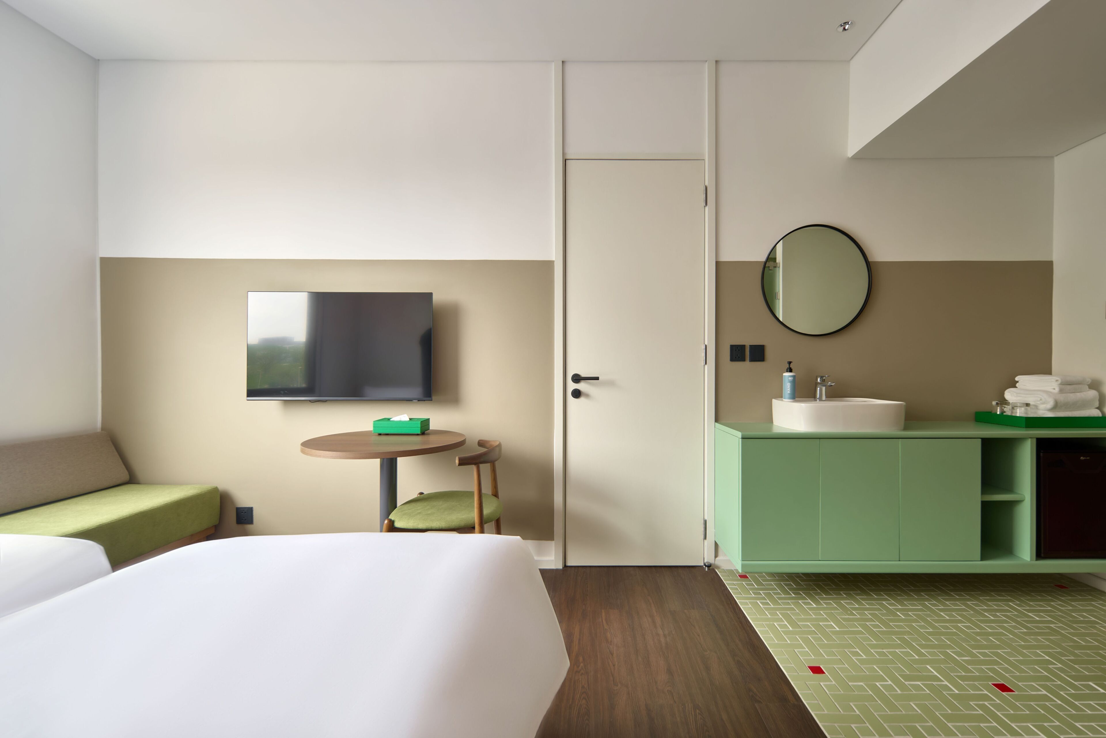 Habitación de hotel con cama, televisión, una pequeña mesa con silla, sofá verde, baño con lavamanos y espejo, y un armario verde.