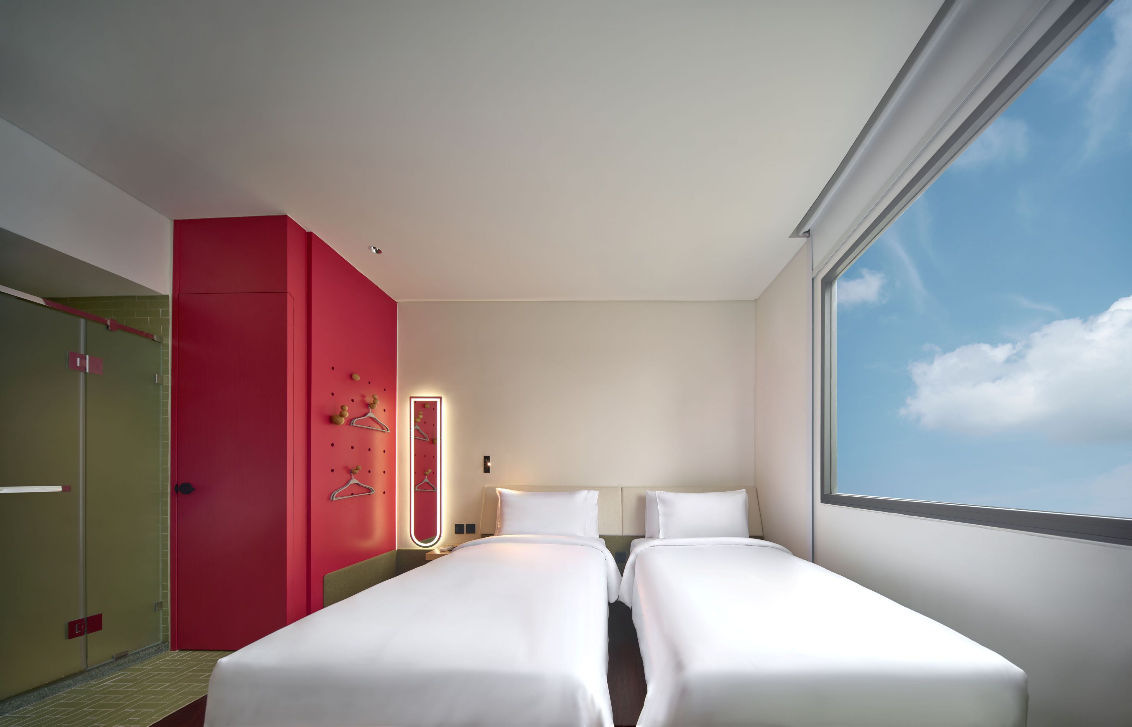 A hotel room with two beds, a red wall, a large window, and a view of the sky.