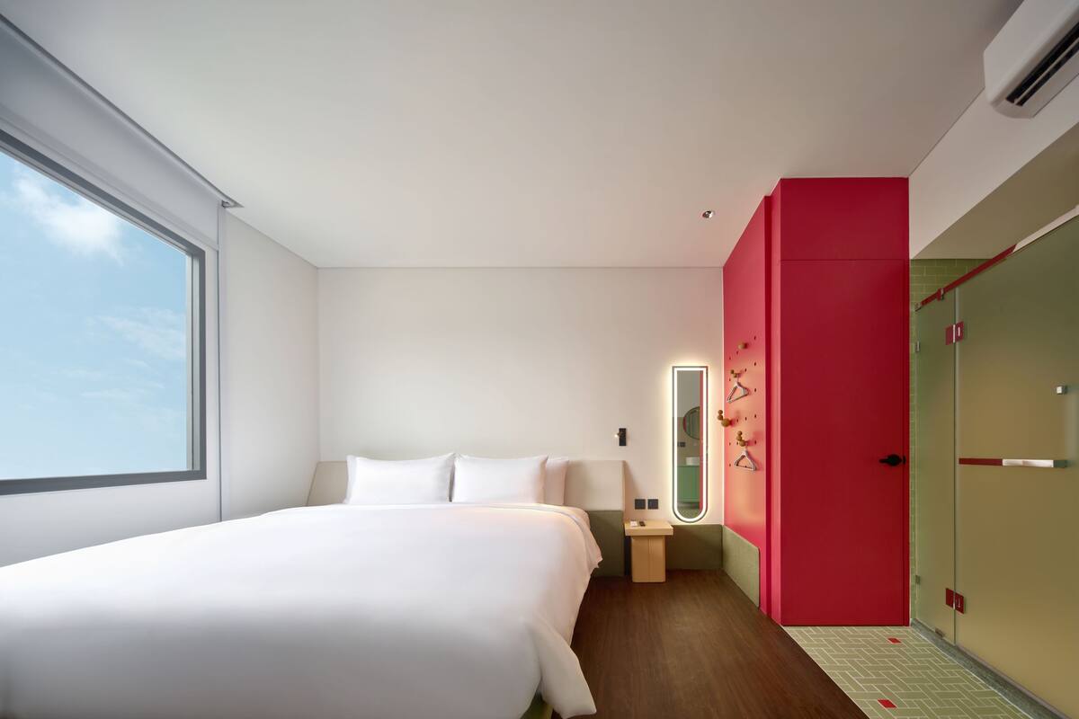Une chambre d’hôtel avec un grand lit, une table de chevet, un miroir, une armoire rouge et une fenêtre.