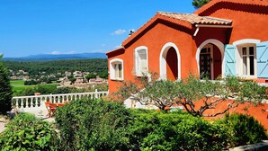 Un edificio rosso con archi bianchi e persiane blu, affacciato su un villaggio e montagne.
