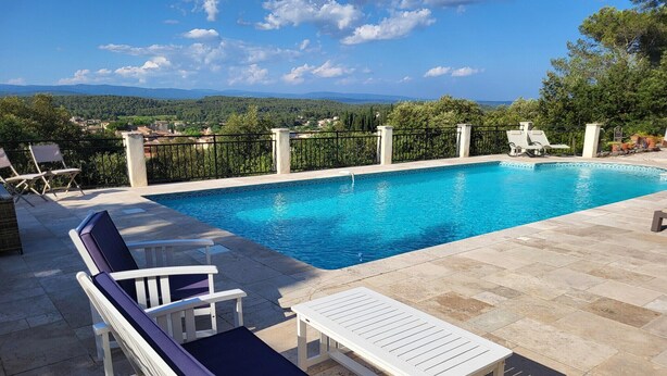 Una piscina rettangolare con acqua cristallina, circondata da un solarium piastrellato. Sul solarium ci sono lettini e un tavolo, e sullo sfondo si vede un paesaggio verde con alberi e colline.