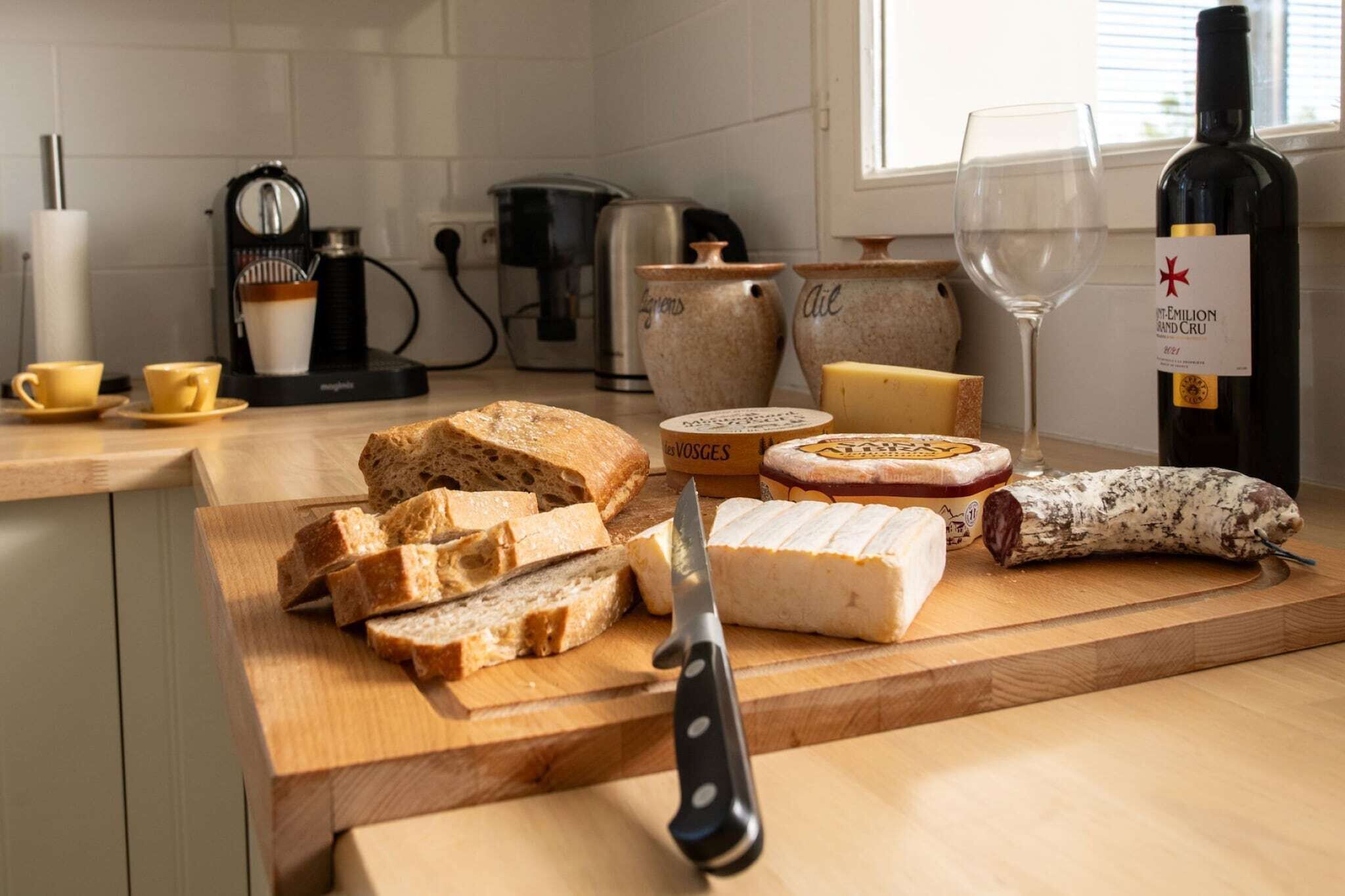 A wooden cutting board with various cheeses, a knife, and a bottle of wine on a kitchen counter.
