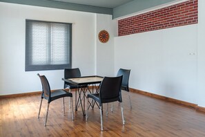 A room with a wooden floor, a round table with chairs, a window with blinds, a dartboard, and a brick wall.