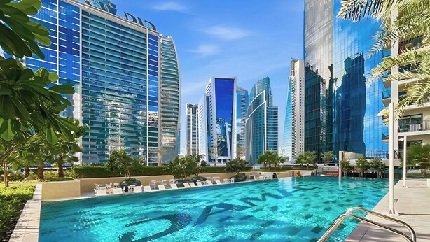 A swimming pool with clear blue water, surrounded by modern high-rise buildings and palm trees. - Canal View 1BR Near Business Bay Metro (Dubai)