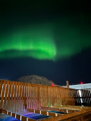 Eine Holzterrasse mit Sitzgelegenheiten, ein Berg im Hintergrund und das Nordlicht am Himmel.