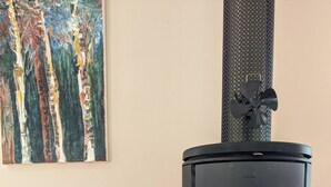A wood-burning stove with a glass door, a log pile, and a painting on the wall. - Mountain Blue Guest house (Golden Valley)