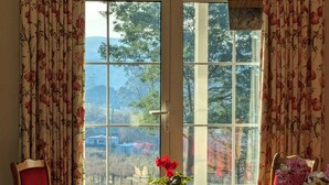 A dining area with a table set for four, a vase of flowers, and a view of a mountainous landscape through large windows. - Mountain Blue Guest house (Golden Valley)