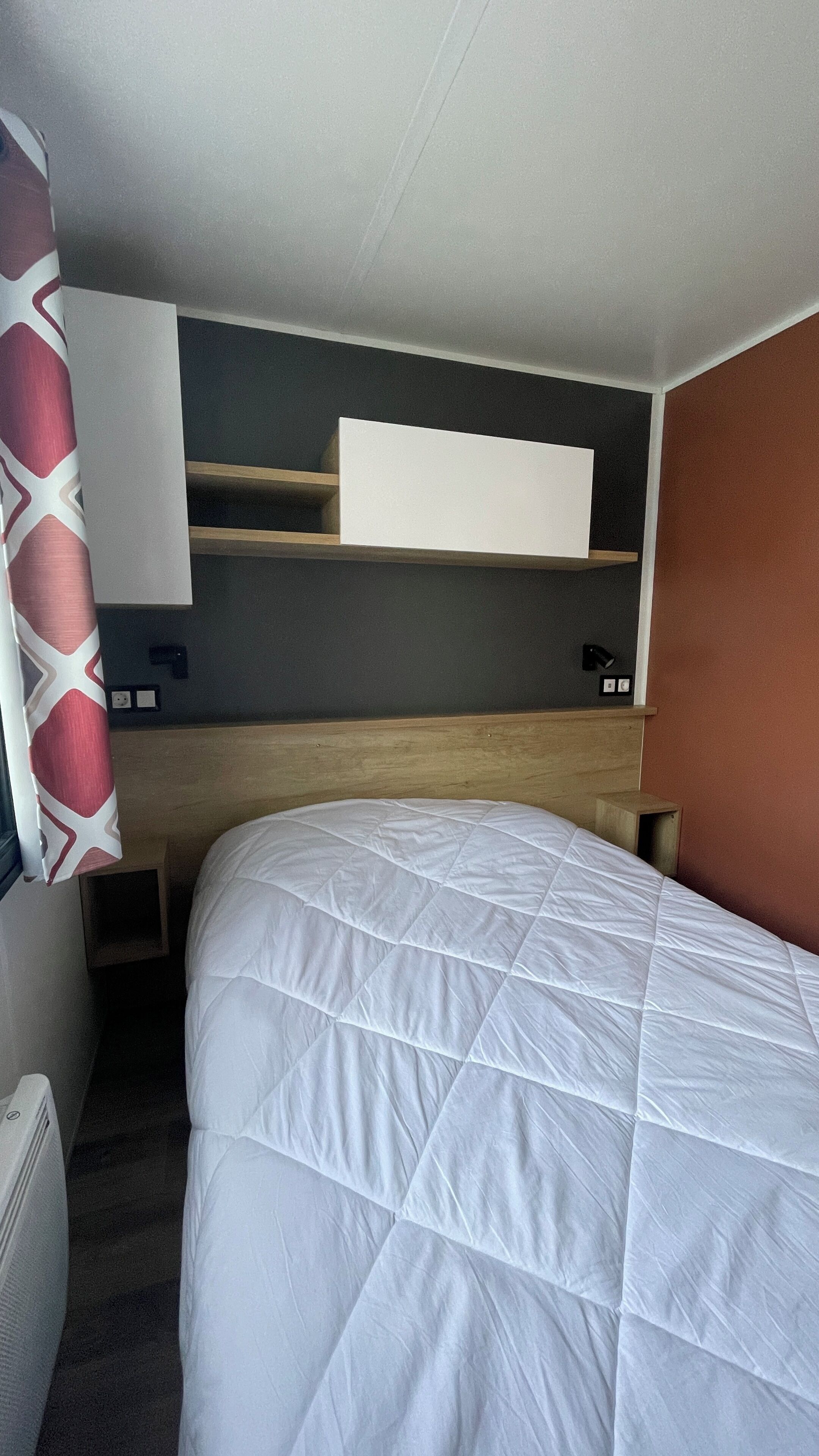 Une chambre d’hôtel avec un lit, une tête de lit en bois, un mur blanc et une étagère au-dessus du lit.