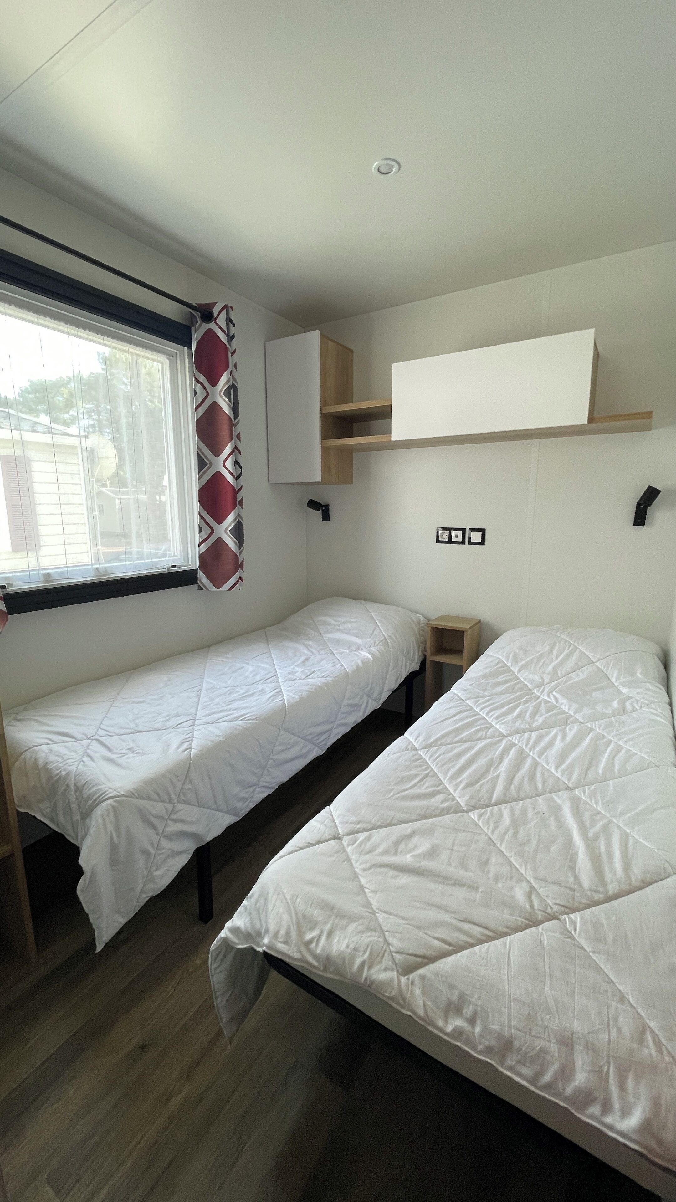 A small, clean room with two single beds, a window with curtains, a shelf with a mirror, and a wall-mounted light.