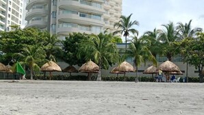 Een strand met rieten parasols, palmbomen en een gebouw op de achtergrond.