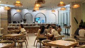 Een restaurant met houten tafels en stoelen, hangende rieten lampen en een keuken in de achtergrond.