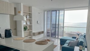 Een moderne woonruimte met een keukenhoek, een eettafel en een zithoek. Een groot raam biedt uitzicht op zee.