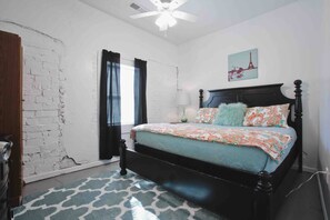 A bedroom with a bed, a ceiling fan, a window with curtains, and a wall with a partial brick wall.