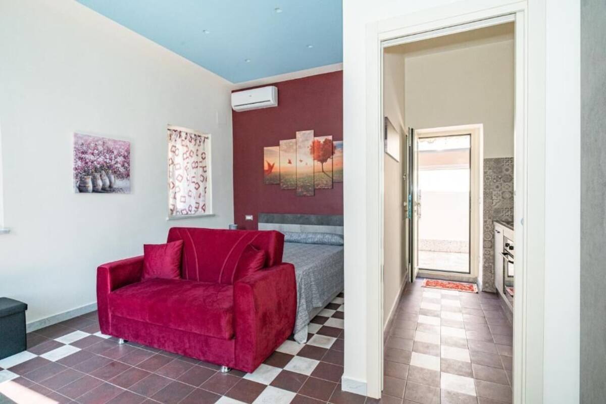 A room with a red sofa, a bed, and a checkered floor. There are wall art pieces and an air conditioning unit.