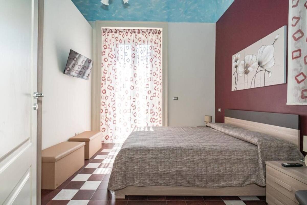 A bedroom with a checkered floor, a bed with a grey blanket, a bedside table, and a wall with a floral painting.