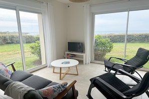A room with a sofa, two lounge chairs, a small table, a TV on a stand, and large windows with a view of the sea.