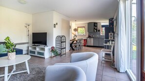 Uma sala de estar moderna com uma televisão de tela plana, sofás brancos, uma mesa de jantar e uma cozinha ao fundo.