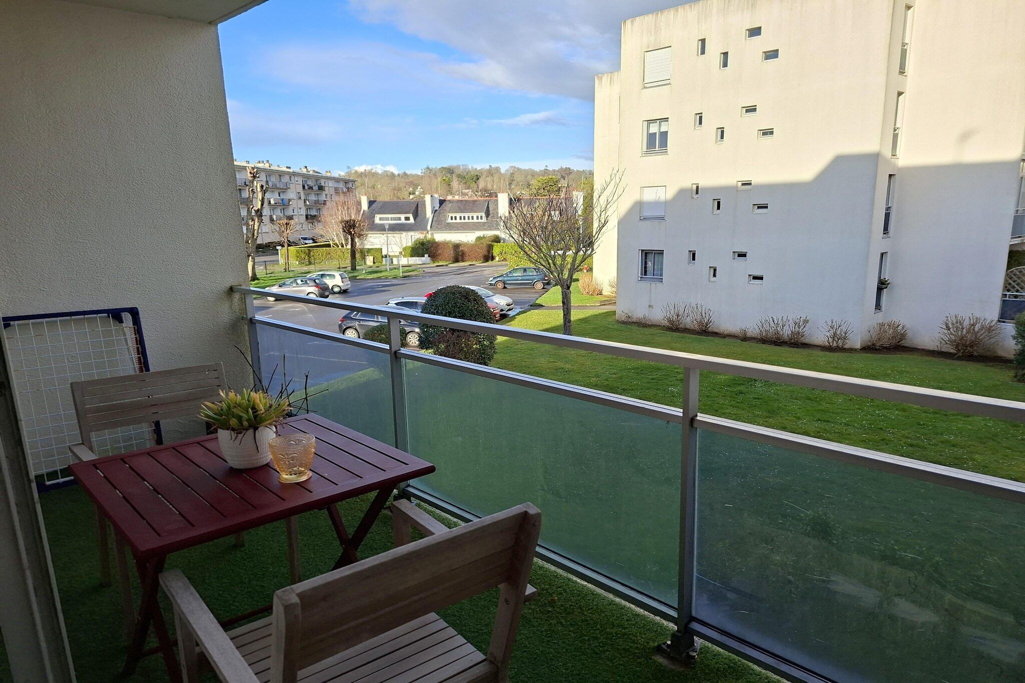 A balcony with a table and chairs, a potted plant, and a view of a street with parked cars and a building in the background.