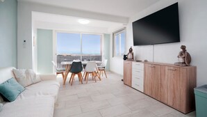 Una sala de estar moderna con un sofá, una mesa de comedor con sillas, un televisor montado en la pared y vistas al mar a través de grandes ventanales.
