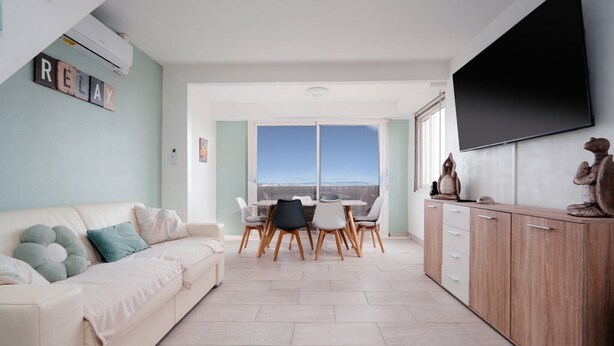 Una sala de estar con un sofá, una mesa de comedor con sillas, un televisor montado en la pared y un ventanal con vistas.
