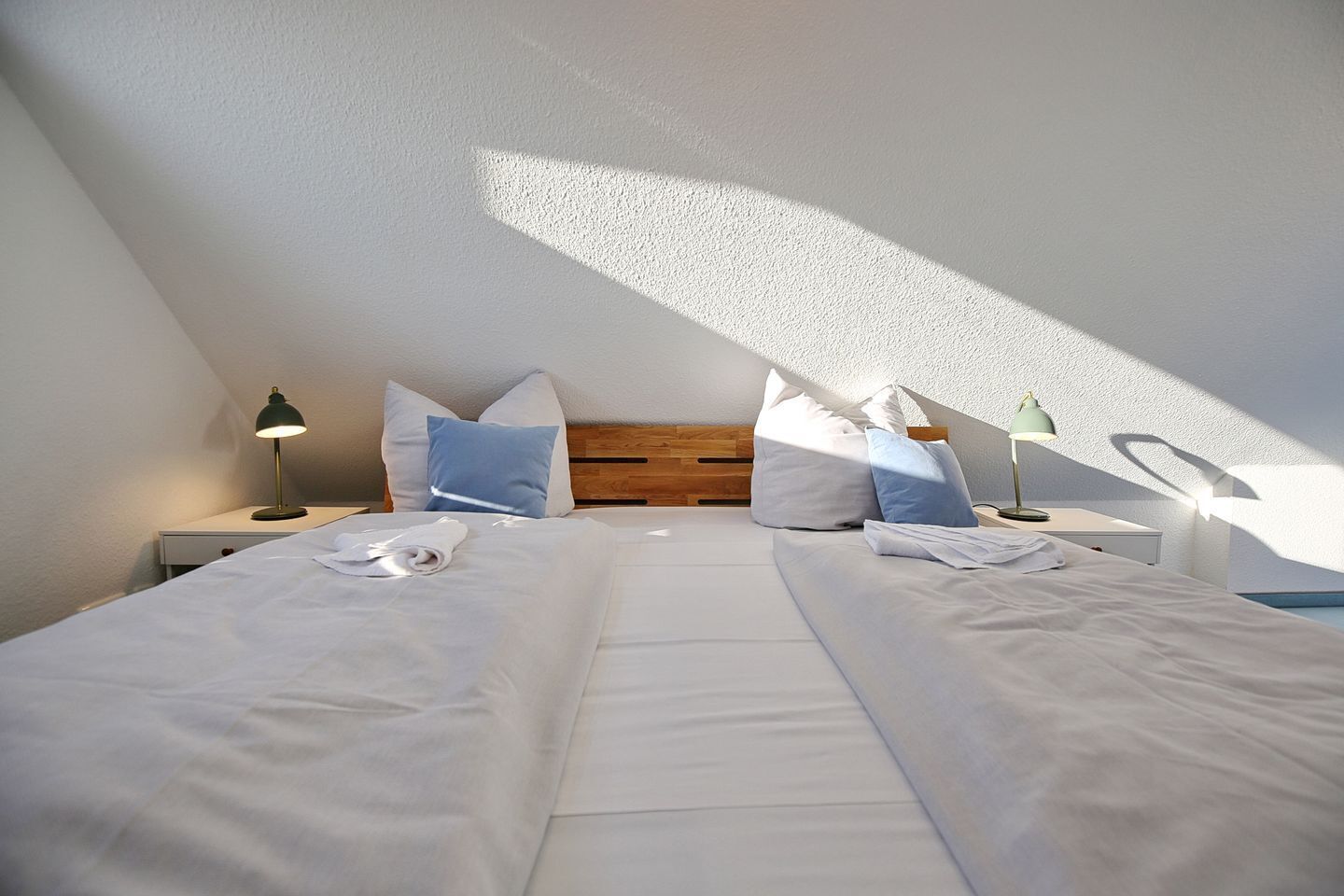 A bedroom with a sloped ceiling, a wooden headboard, two bedside tables with lamps, and a neatly made bed with pillows and a folded towel.