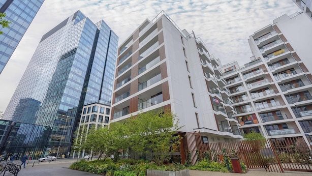 Ein moderner Gebäudekomplex in der Stadt mit mehreren Hochhäusern, einem Fußgängerweg und einigen Grünflächen.
