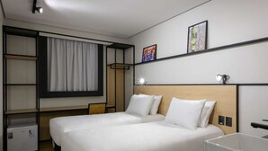 Habitación de hotel con dos camas, un escritorio, un refrigerador pequeño y una ventana con cortinas.