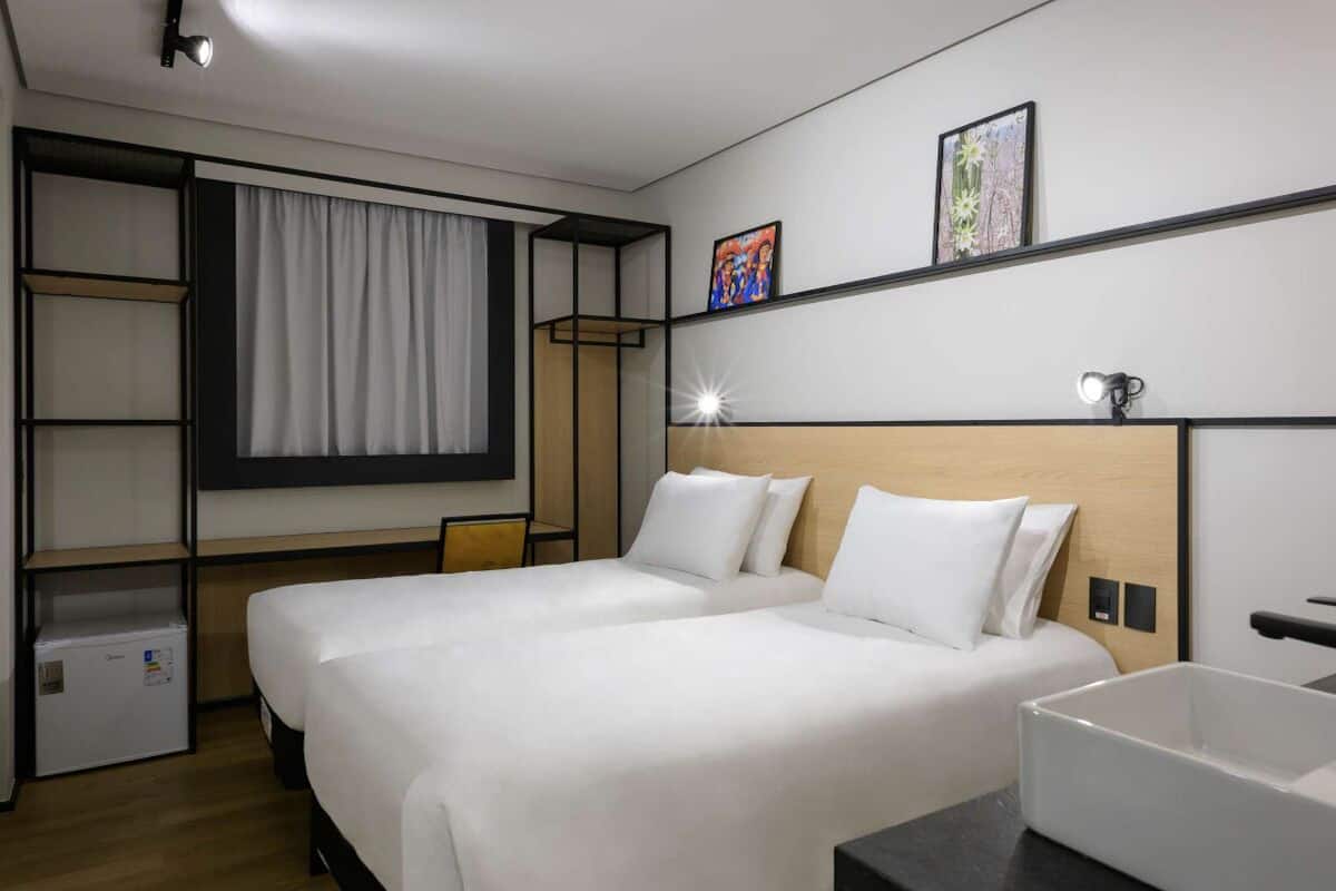 Habitación de hotel con dos camas, un escritorio, un refrigerador pequeño y una ventana con cortinas.