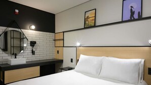 Habitación de hotel con cama, mesita de noche, espejo, lavamanos y luz empotrada en la pared.