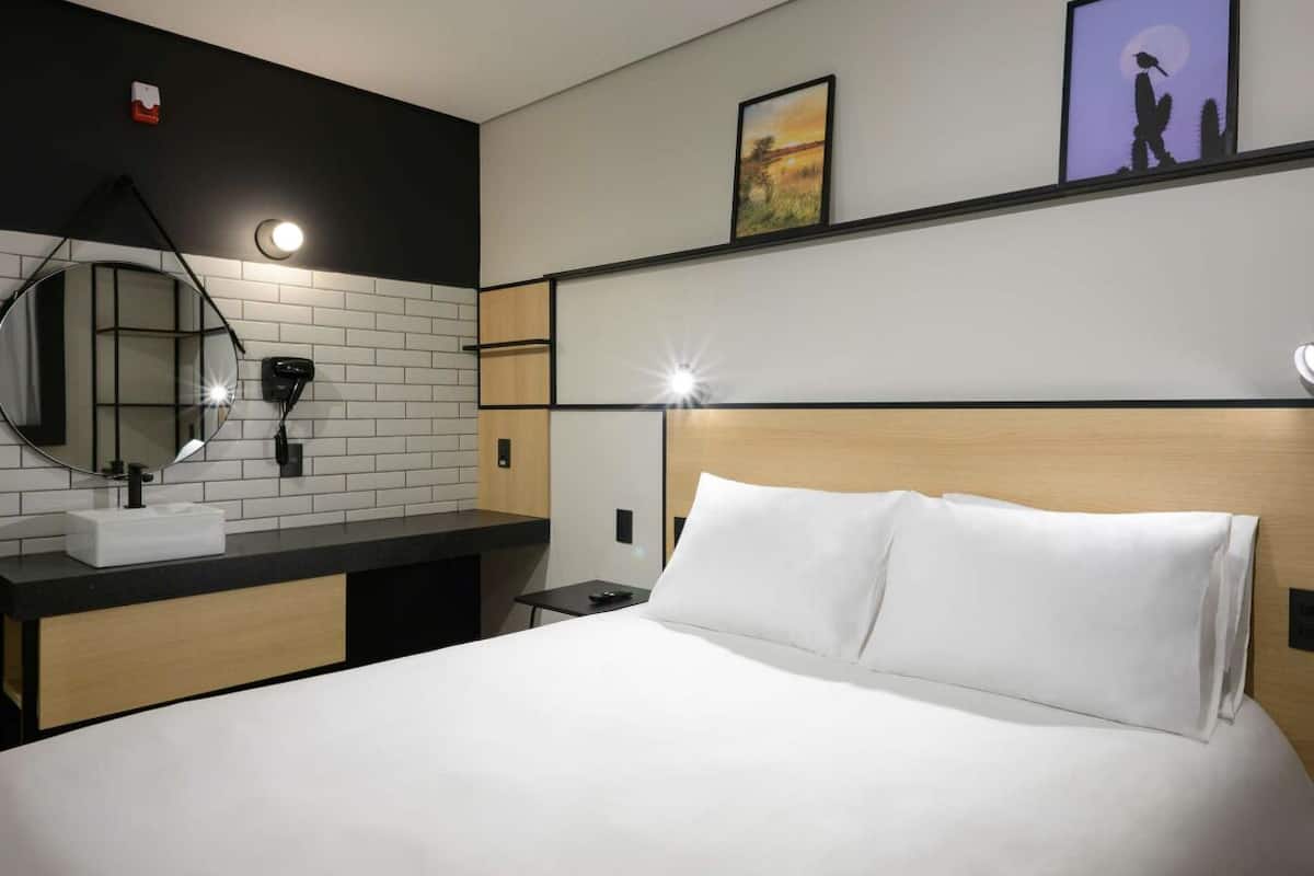 Habitación de hotel con cama, mesita de noche, espejo, lavamanos y luz empotrada en la pared.