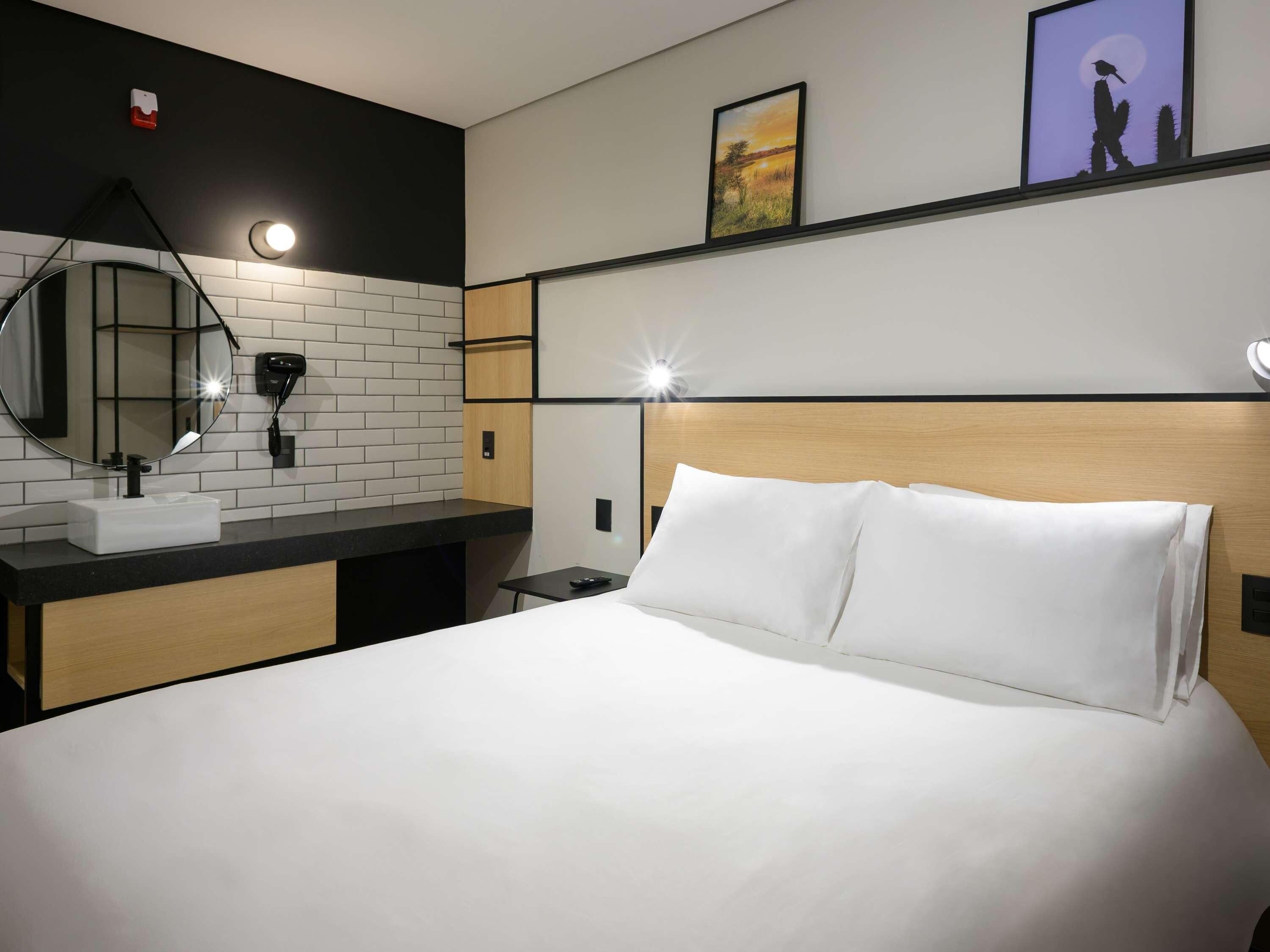 Quarto de hotel com uma cama, um espelho redondo, uma pia e uma mesa de cabeceira.