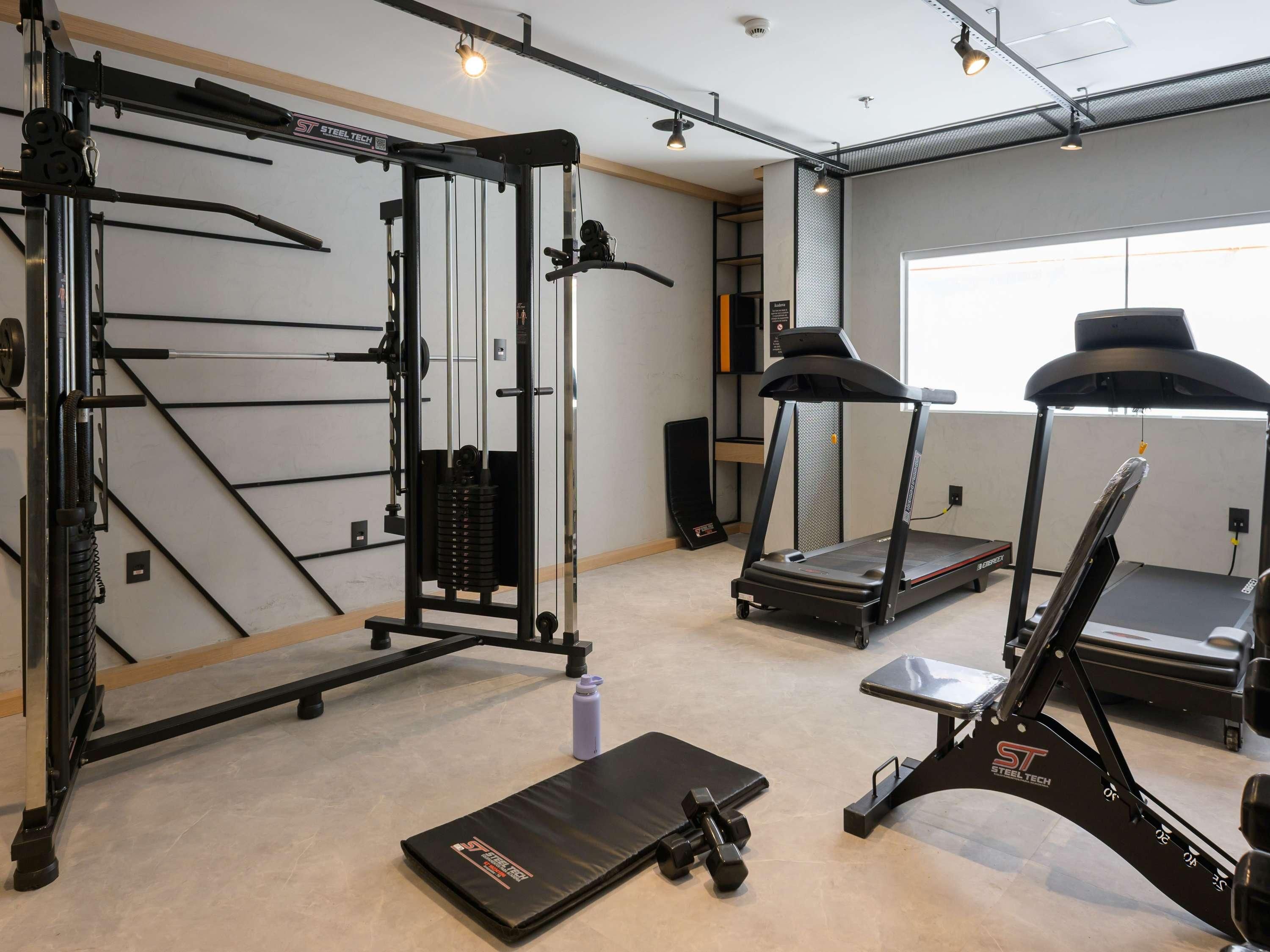 Uma academia com barra de exercícios, banco de musculação, esteira e aparelho de supino.