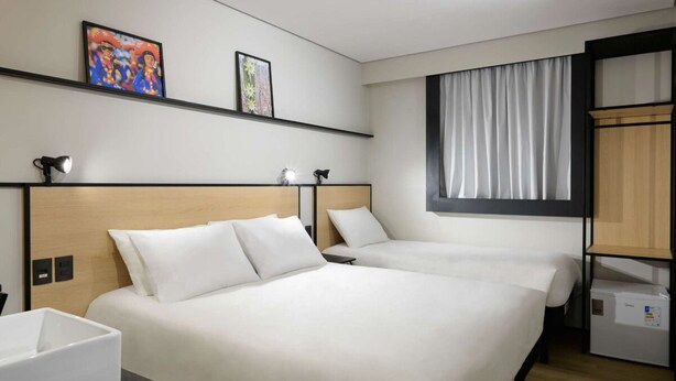 Habitación de hotel con dos camas, una mesita de noche, un armario pequeño y una ventana con cortinas.