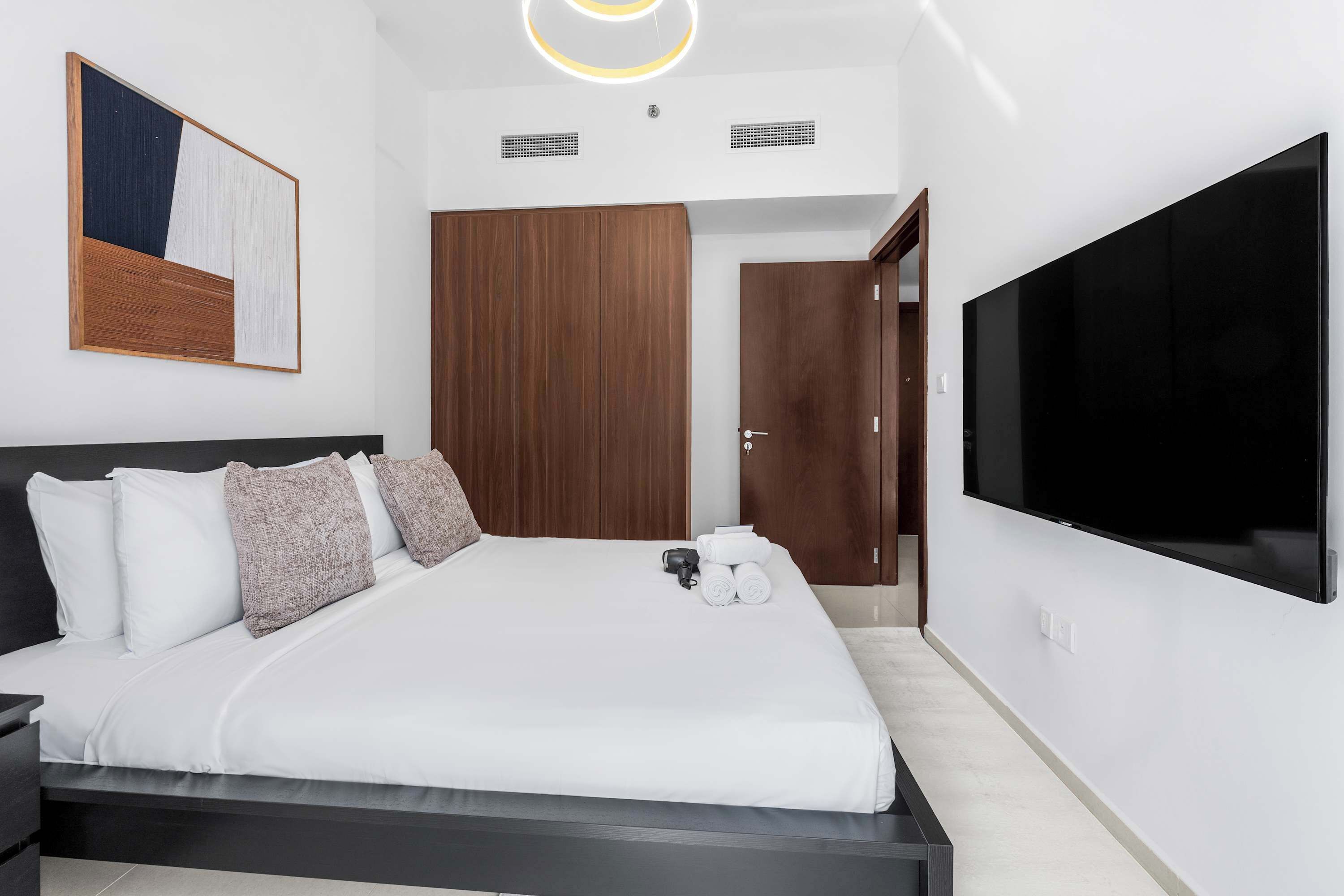 A modern hotel room with a large bed, a flat-screen TV, a wooden wardrobe, and a painting on the wall.