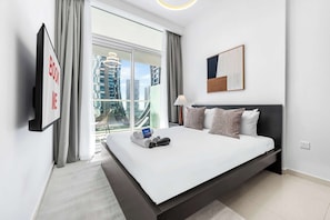 A modern hotel room with a large bed, a TV mounted on the wall, a city view, and a framed artwork above the bed. - StayShort - Cozy & Modern 1BR in Business Bay that Sleeps 4! (Damac)