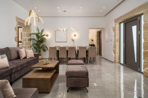 Ein modernes Wohnzimmer mit Sofa, Hocker, Couchtisch, Esstisch, Stühlen, Pendelleuchten und einer Topfpflanze.