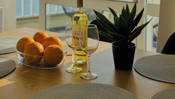 Una mesa con una botella de vino, copas y naranjas, con vista al mar a través de una puerta de balcón.