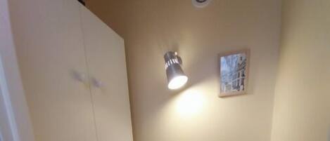 A small bathroom with a toilet, a shelf with toiletries, a wall-mounted light, and a vent.
