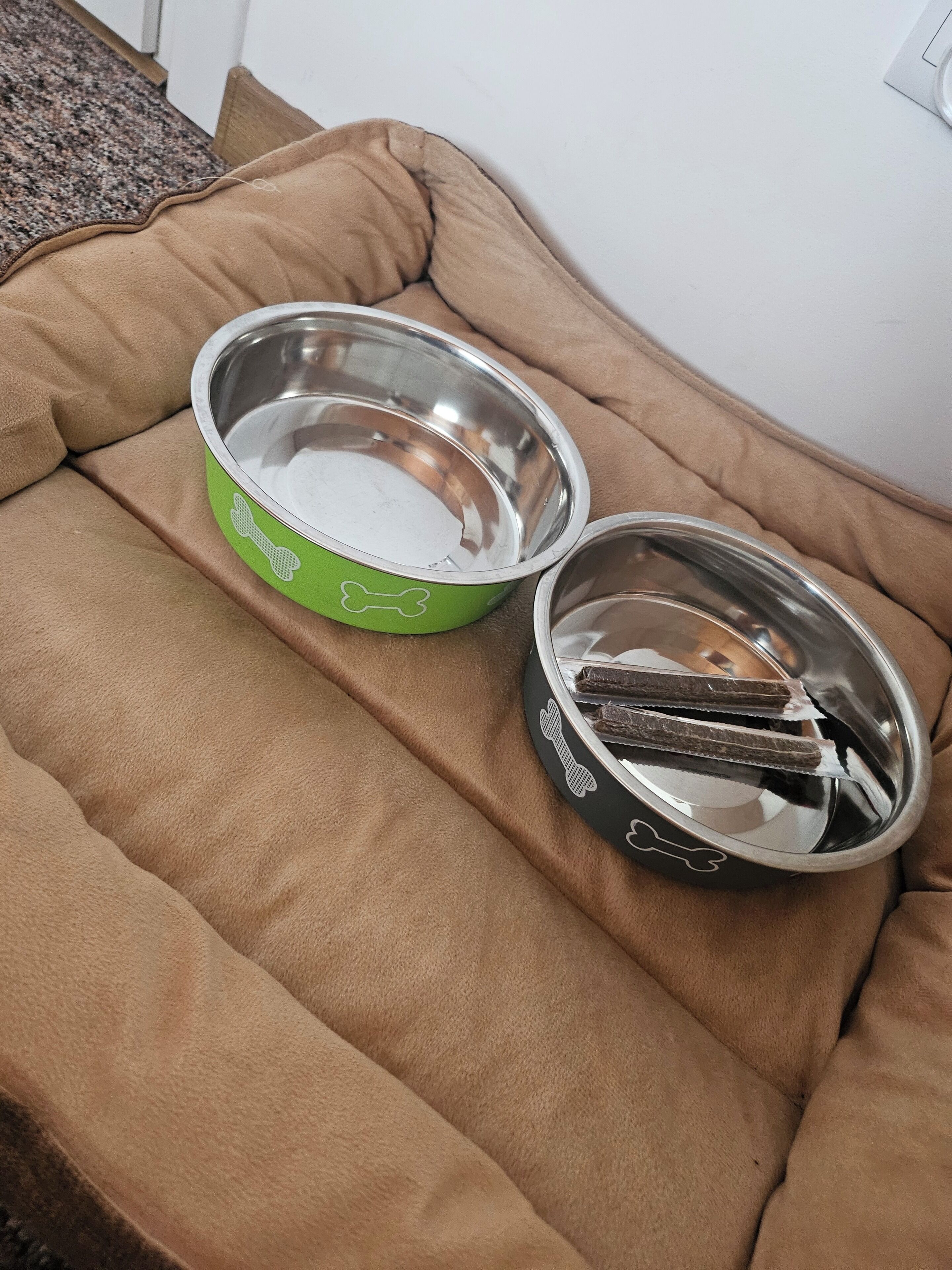 A pet bed with two metal bowls, one green and one black, containing food.