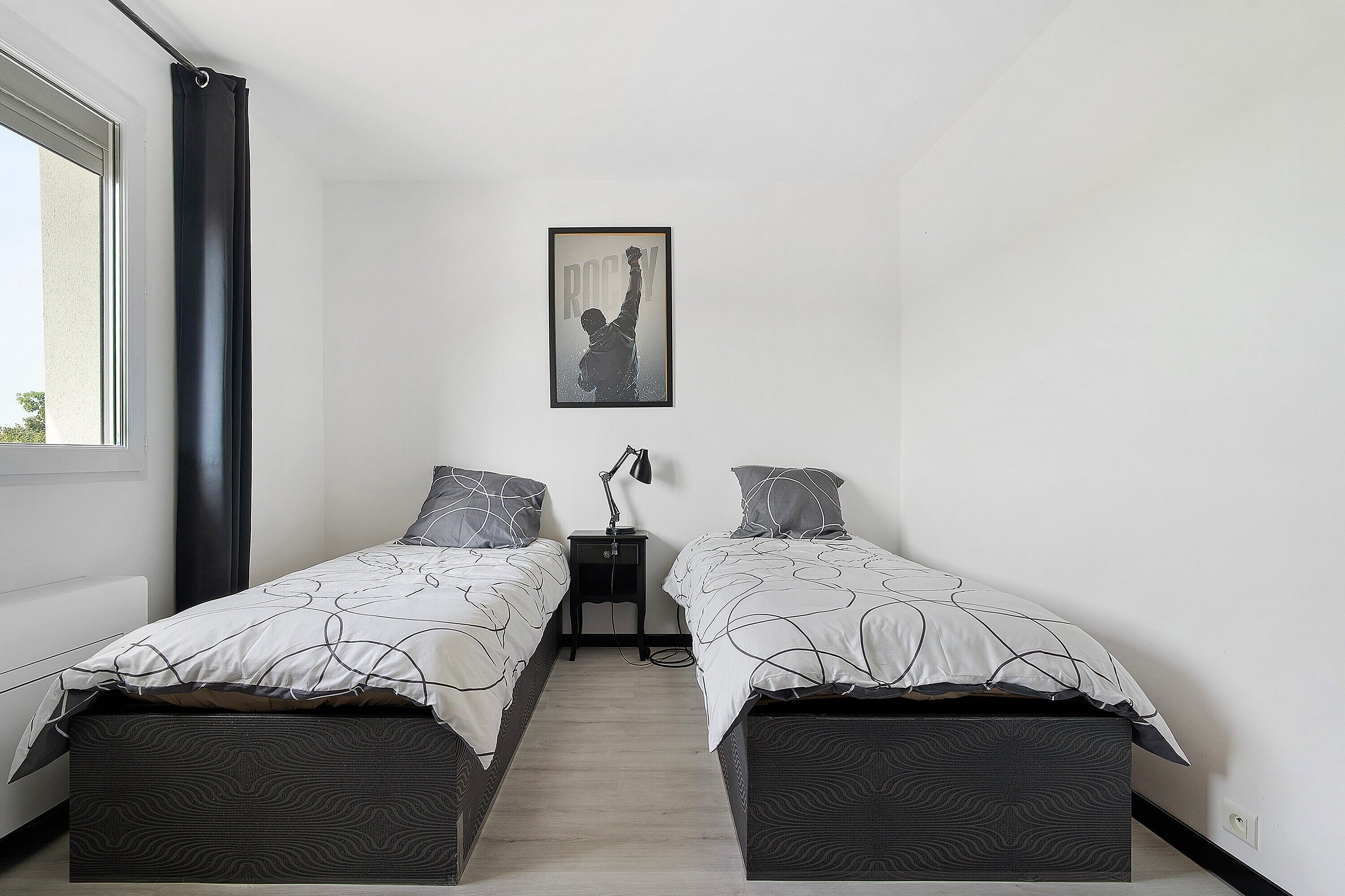 Une chambre avec deux lits, une table de chevet, un tableau encadré au mur et une fenêtre avec des rideaux.