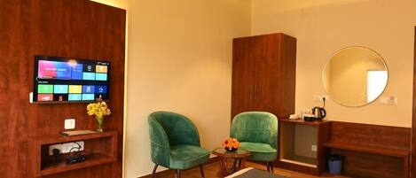 A modern hotel room with a wooden panel wall, a flat-screen TV mounted above a shelf, two green armchairs, a small round table with a vase of flowers, a round mirror, and a bed with a gray quilt.