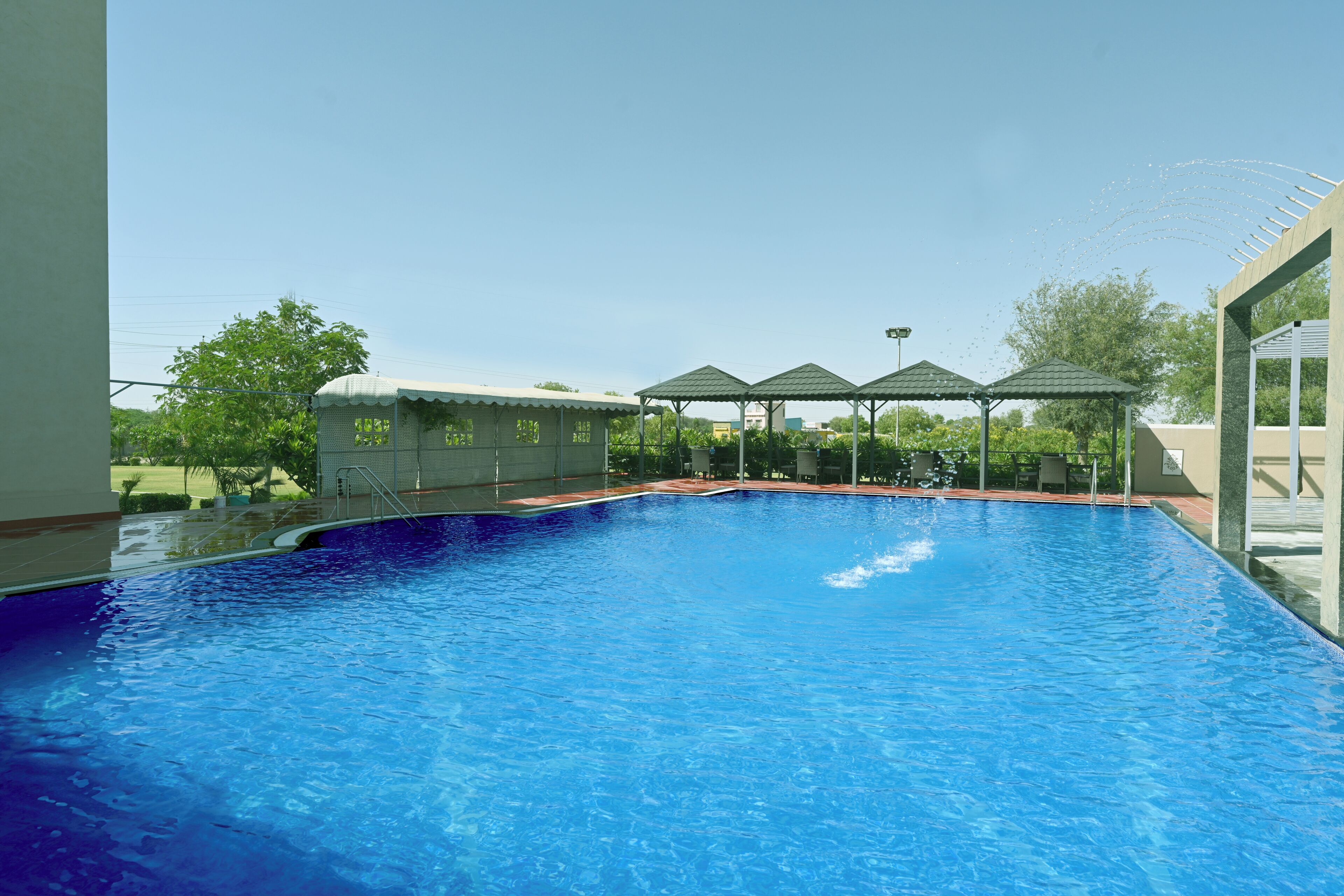 A swimming pool with a clear blue water, surrounded by a covered area with a roof, and a small building with a dome roof in the background.